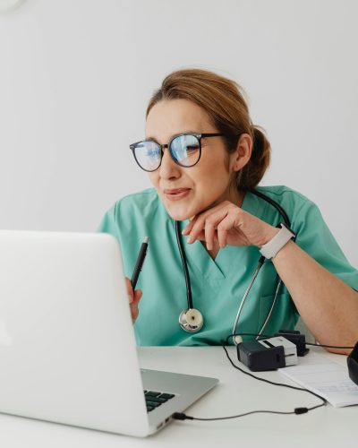 A healthcare professional wearing scrubs and eyeglasses using a laptop with a stethoscope around the neck.