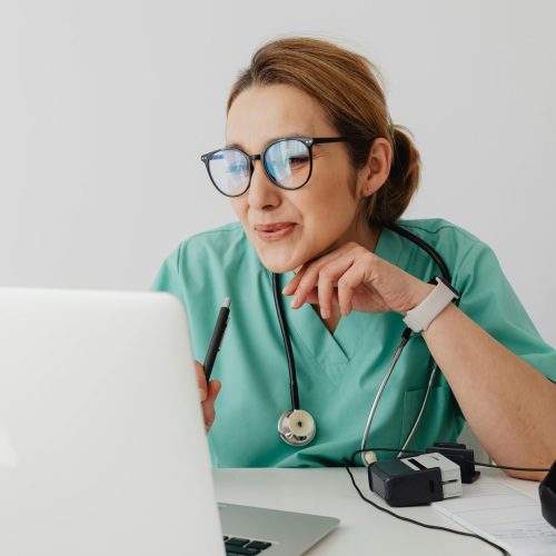 A healthcare professional wearing scrubs and eyeglasses using a laptop with a stethoscope around the neck.